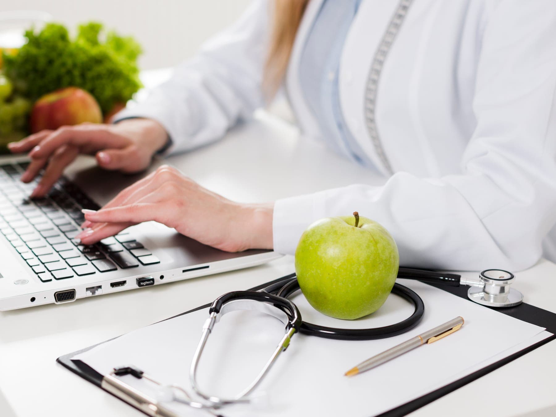 Nutricionista escribiendo en el computador junto a una manzana y un estetoscopio